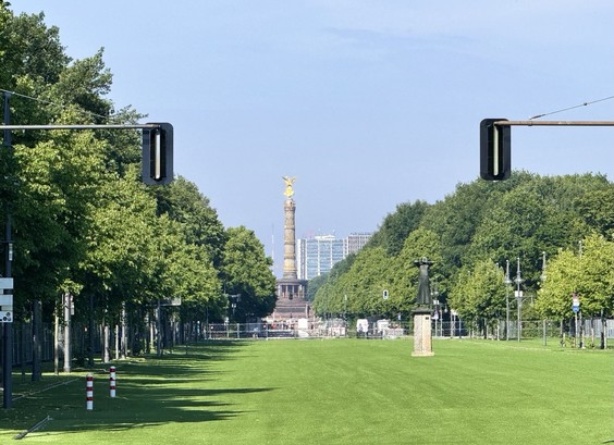 View of a tree-lined avenue leading to a tall monument topped with a golden statue, with traffic lights and other sculptures visible in the foreground.