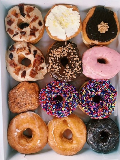 An open box with 12 donuts with different colourful toppings seen from above, looking tentatively at the viewer.