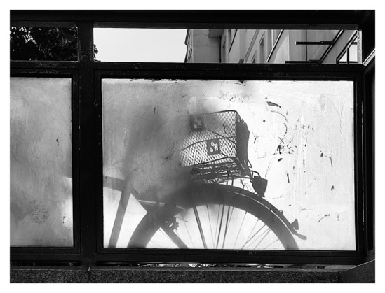 Schwarz-Weiß-Foto. Blick durch ein Geländer, das aus großen Milchglasscheiben besteht. Dahinter steht ein Fahrrad, dessen hinterer Teil sich als sehr deutlicher Schatten auf dem Milchglas abbildet.