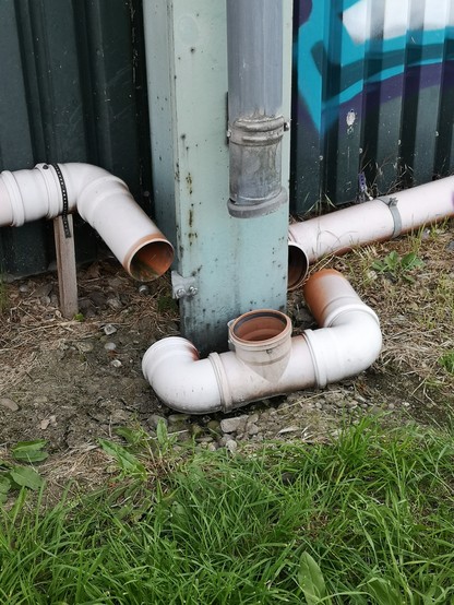 Drei Wasserrohre an einem Gebäude treffen an einem Pfeiler ohne Verbindung offen aufeinander - das passende (?) Verbindungsstück liegt nutzlos davor 