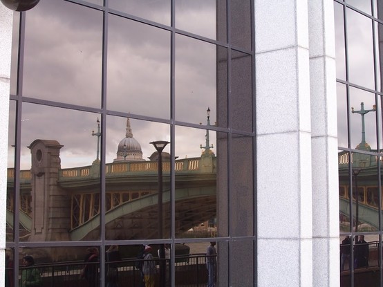 a modern facade in glass, aluminium and white stone with the reflection of some people at the south bank of the river Thames, the arcs of Southwark bridge in green and yellow and behind the roof of St. Paul's Cathedral.