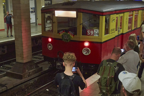 Eine alte gelb-rote S-Bahn steht auf einem Gleis. Vorne ist eine 100 angebracht sowie die Jahreszahlen 1924 und 2924 auf Deutschlandflaggen. Darum herum stehen Menschen, die die Bahn mit ihren Handys fotografieren.