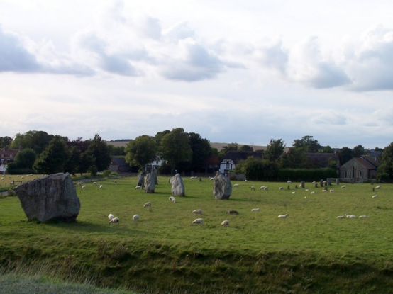 Sheep between standing stones on a green meadow under a cloudy sky. In the background, trees and an English village with small white houses and a chapel built with grey stones.  