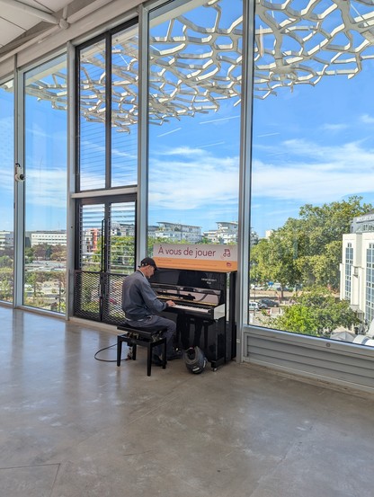 Jemand spielt im Bahnhof an einem öffentlichen Klavier. das Klavier steht vor einem großen Fenster mit Stadtkulisse und blauem Himmel.