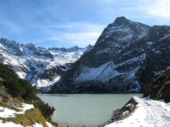 A lake in the mountains, a footpath covered with snow to the right. The mountains behind are partly covered with snow, too. 