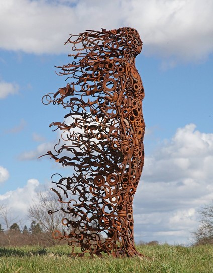 A female statue made from scrap metal standing on a meadow. 

The front part is mainly made from round pieces, like holes where the wind can blow through. 

The back part is from longer pieces, like hair blowing in the wind. I  a way, it seems like she is disintegrating, neing nlown away and yet standing firm.